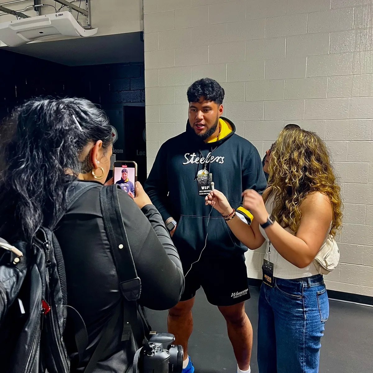 PMN+ team filming a post-game interview with a player in a stadium locker-room corridor